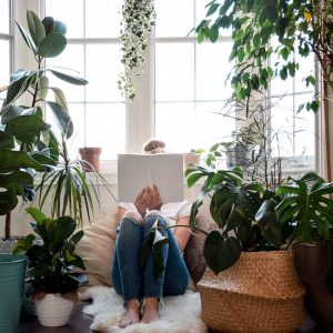 Young girl sitting by a window with houseplants reading a book covering her face